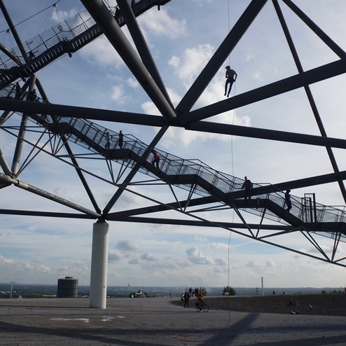 Menschen steigen Treppen und hangeln an einem Stahlgerüst unter einem wolkenverhangenen Himmel.