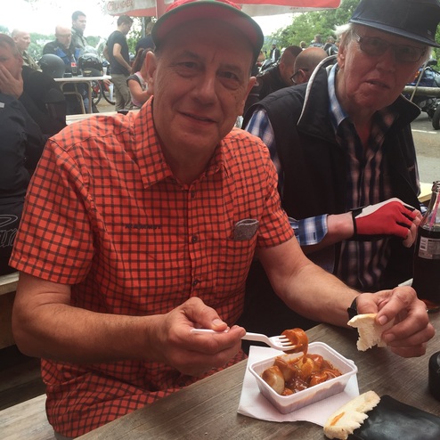 Lächelnder Mann mit Kappe hält eine Gabel und isst Wurst mit Soße aus einer Schale, während ein weiterer Mann daneben sitzt.