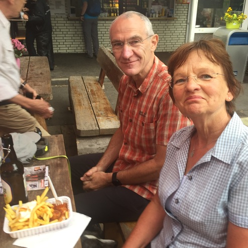 Paar sitzt an einem Holztisch, teilt sich Pommes und Currywurst, im Hintergrund Gäste und ein Imbissstand.