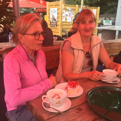Zwei Frauen sitzen an einem Holztablett im Freien und genießen Kaffee und Kuchen. Pflanzen und eine weitere Person im Hintergrund.