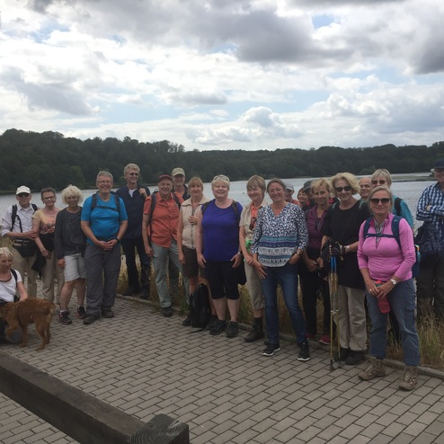 Gruppe von Wanderern am Ufer eines Sees, umgeben von Bäumen und bewölkem Himmel. Hund sitzt vor einer Person.