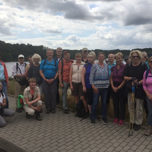 Gruppe von Wanderern steht am Ufer eines Sees, umgeben von Bäumen und bewölktem Himmel. Einige halten Wanderstöcke.