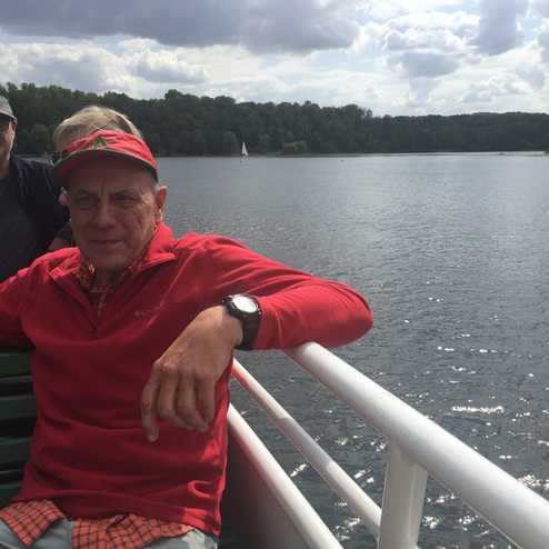 Mann in rot-gestreifter Jacke sitzt an Bord eines Schiffs, Blick auf einen ruhigen See und umgebende Wälder.