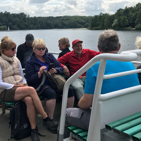 Gruppe von Menschen auf einem Boot, einige unterhalten sich, andere nutzen Handys, im Hintergrund ein See und Bäume.