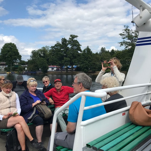 Eine Gruppe von Touristen sitzt auf einem Boot, einige halten Handys, während die Umgebung am Wasser zu sehen ist.