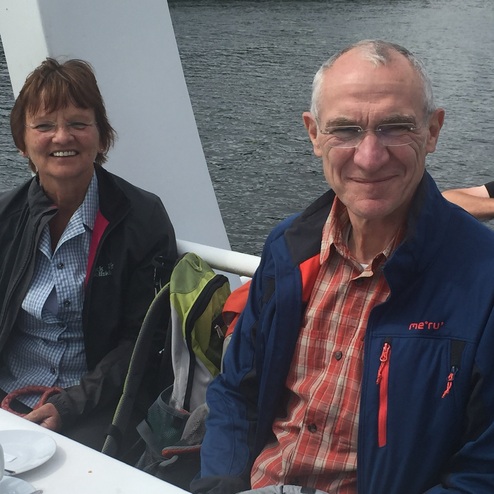 Paar sitzt auf einem Boot, lächelt in die Kamera, im Hintergrund Wasser und eine weitere Person.