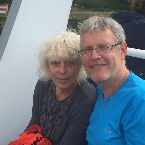 Paar mit blonden Haaren sitzt auf einem Boot, im Hintergrund Wasser und grüne Landschaft. Sie lächeln in die Kamera.