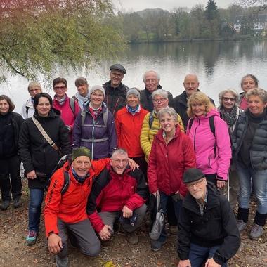 Gruppe von 24 Wanderern steht am Ufer eines ruhigen Sees, umgeben von Bäumen und herbstlicher Landschaft.