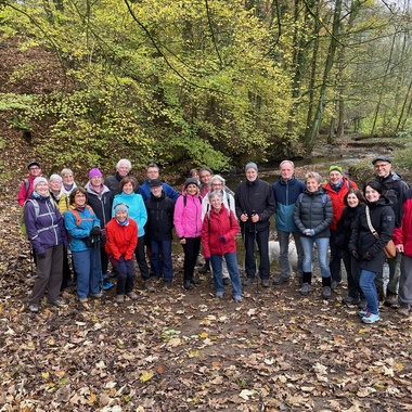 Gruppe von 24 Personen steht in einem herbstlichen Wald mit buntem Laub und einem kleinen Fluss im Hintergrund.