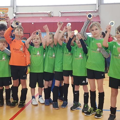 Kinderfußballmannschaft in grünen Trikots zeigt stolz ihre Medaillen nach einem Turnier. Zuschauer im Hintergrund applaudieren.