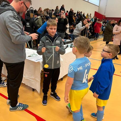 Kinder in Sportkleidung stehen an einem Tisch, während ein Erwachsener einen goldenen Gegenstand präsentiert.