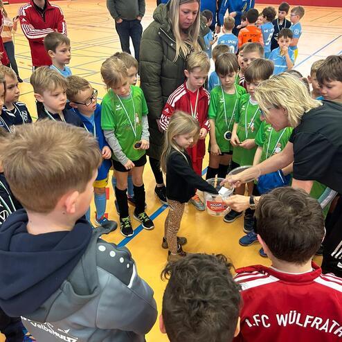 Gruppe von Kindern in Sportshirts versammelt um eine Frau, die Medaillen im Rahmen einer Preisverleihung verteilt.