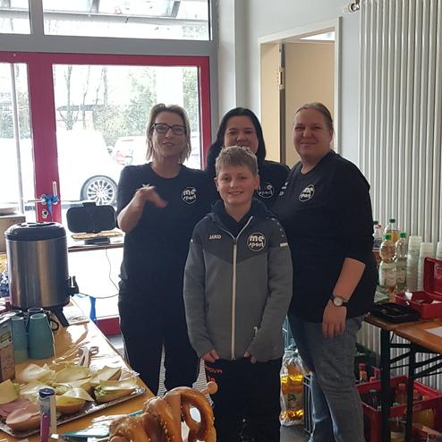 Vier Personen stehen lächelnd vor einem Tisch mit Snacks und Getränken in einem hellen Raum.