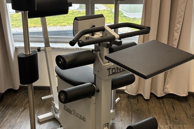 Fitnessgerät mit mehreren gepolsterten Hebeln, platziert in einem Raum mit Fenster und Holzfußboden.