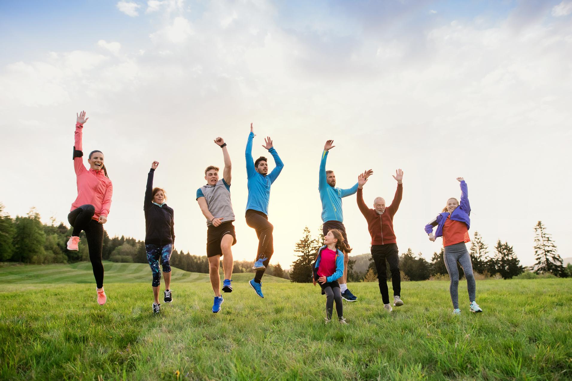Gruppe von sieben Menschen springt fröhlich in einer Wiese, umgeben von Bäumen und einem hellen Himmel.