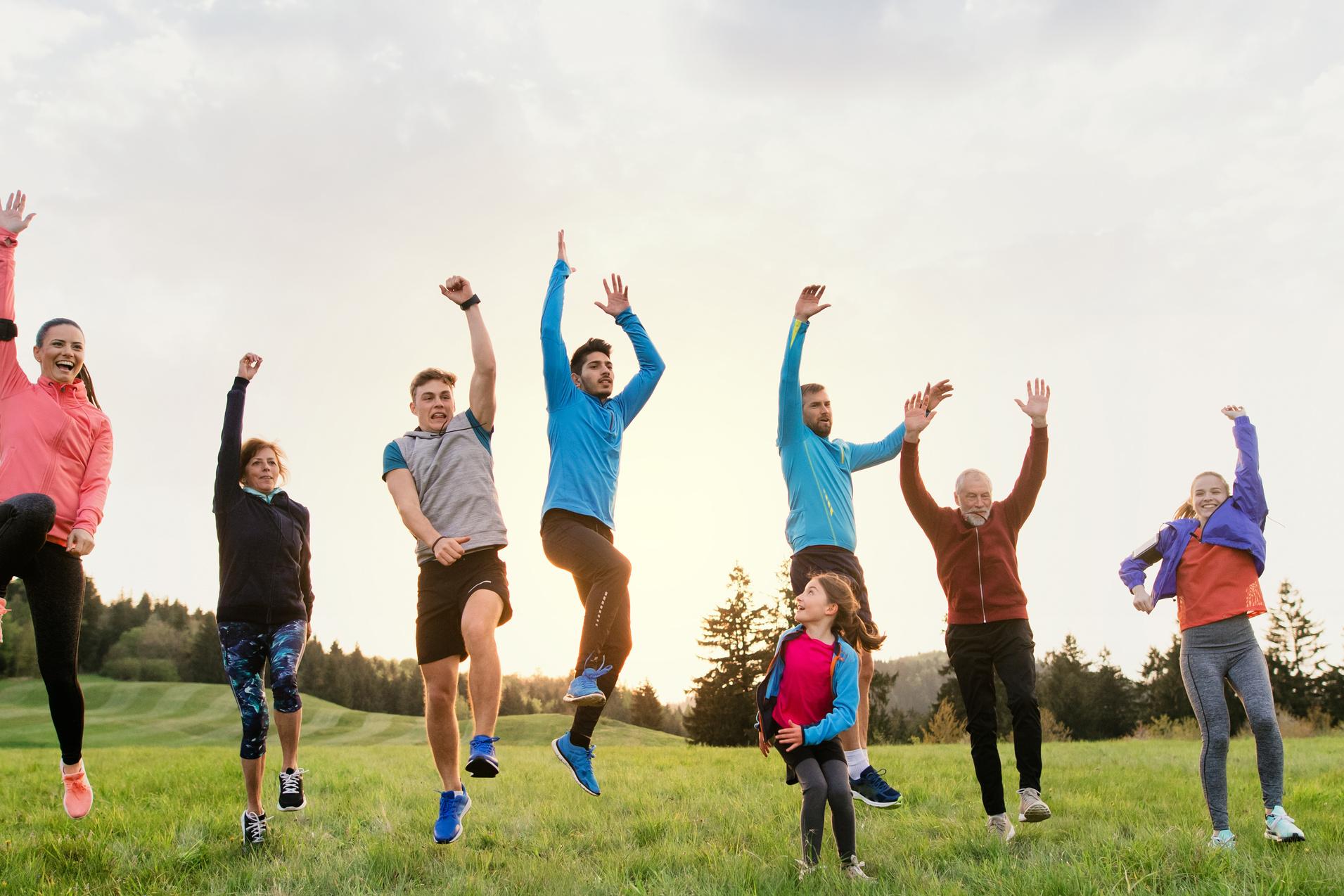 Gruppe von sieben Menschen springt fr&ouml;hlich in einer Wiese, umgeben von B&auml;umen und einem hellen Himmel.
