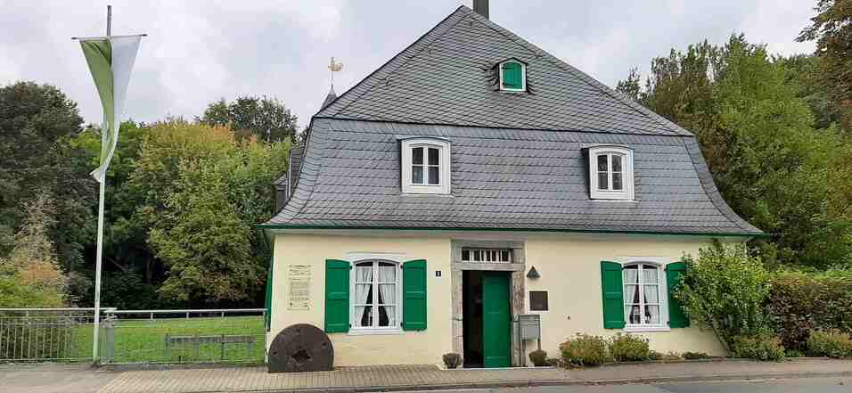 Man sieht das Gebäude der Goldberger Mühle. Die Farben des Hauses sind hellgelb und grau. Links davon steht ein Fahnenmast.