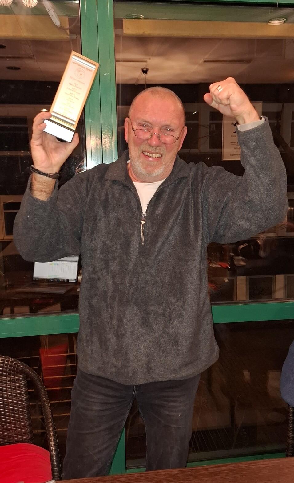 Mann mit Brille hält eine Trophäe hoch und zeigt mit der anderen Hand den Triumph. Hintergrund mit Fenstern und Möbeln.