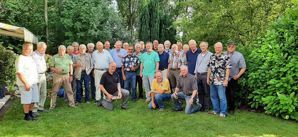 Gruppe von Männern unterschiedlichen Alters posiert im Garten, umgeben von Bäumen und Blumen, einige halten Getränke.