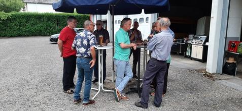 Gruppe von Männern in lässiger Kleidung unter einem Sonnenschirm, stehen um einen Stehtisch und unterhalten sich.