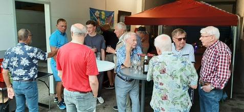 Gruppengespräch von Männern unterschiedlichen Alters bei einer Feier, stehend um einen Stehtisch mit Getränken.