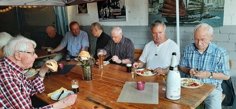 Gruppe älterer Männer sitzt an einem gedeckten Tisch, Essen und Getränke vor sich, in entspannter Atmosphäre.