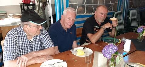 Drei Männer sitzen an einem Tisch, vor ihnen leere Teller und eine Vase mit Blumen. Sie scheinen sich zu unterhalten.