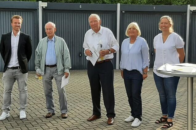 Fünf Personen stehen vor einer Wand mit Garagentoren und lächeln in die Kamera, einige halten Dokumente in der Hand.