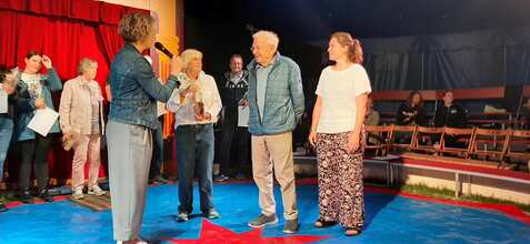 Eine Frau im Jean-Jackett spricht mit einem Mann und einer Frau, die eine Auszeichnung in den Händen halten.