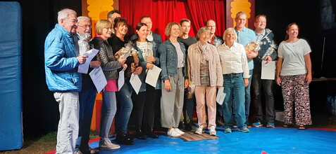 Gruppe von Menschen mit Urkunden und Pokalen auf einer Bühne vor rotem Vorhang, feierliche Atmosphäre.