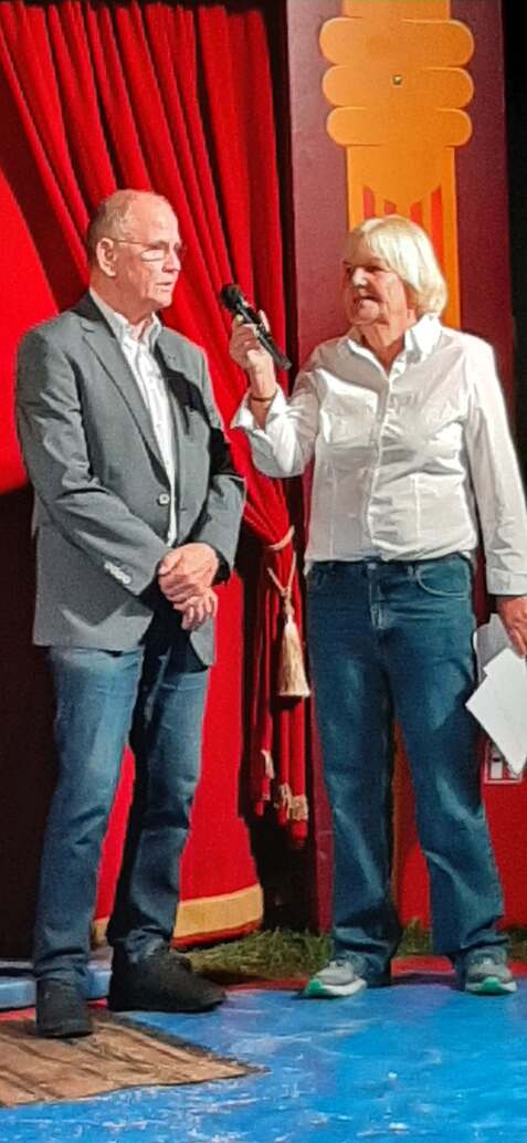 Zwei Personen stehen auf einer Bühne mit rotem Vorhang; eine hält ein Mikrofon und spricht.