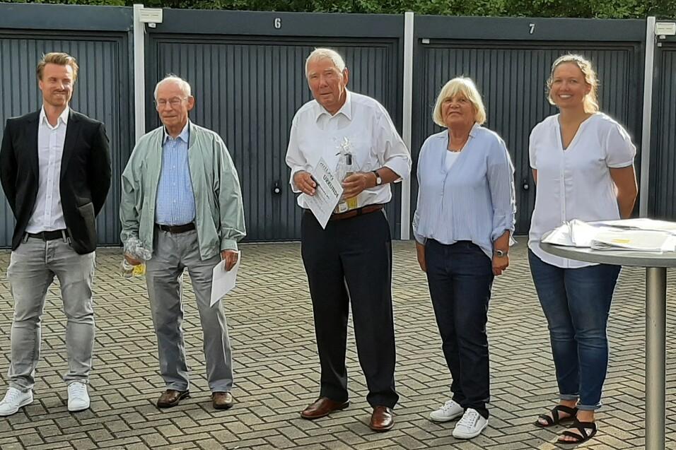 Fünf Personen stehen vor einer Wand mit Garagentoren und lächeln in die Kamera, einige halten Dokumente in der Hand.