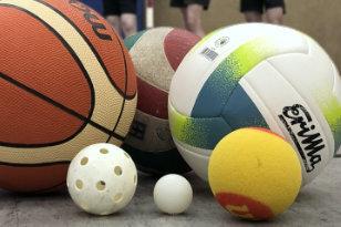 Vielfältige Sportbälle, darunter Basketball, Volleyball und Tischtennisball, liegen auf dem Boden einer Sporthalle.
