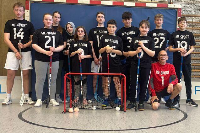 Gruppe von Jugendlichen in Sportshirts mit Bodenballschlägern vor einem roten Tor in einer Sporthalle.