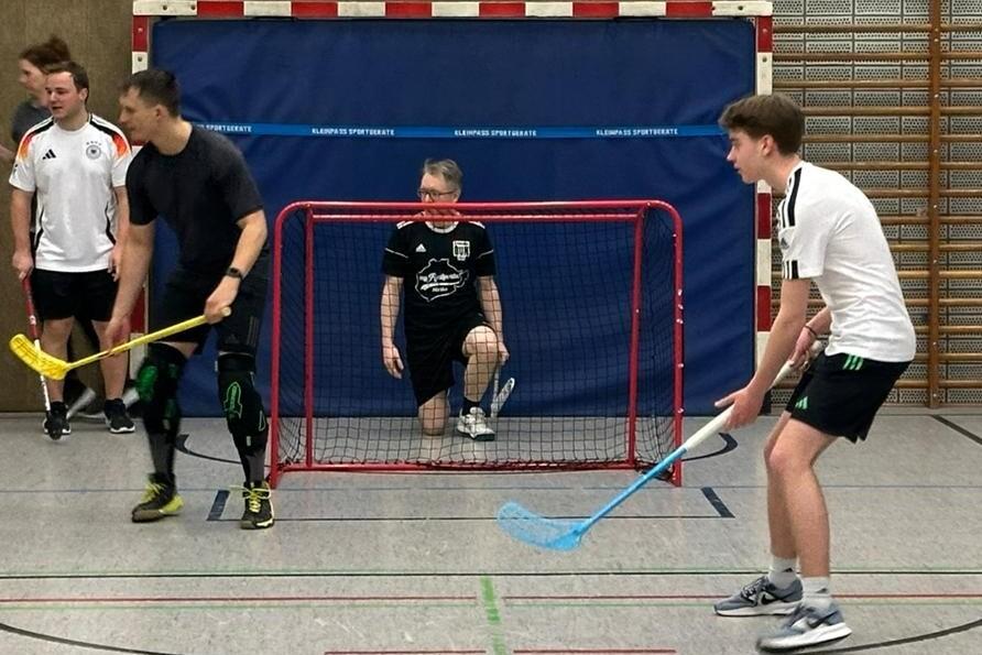 Fünf Personen spielen Floorball in einer Sporthalle, ein Torwart sitzt im Tor, andere halten Schläger in der Hand.