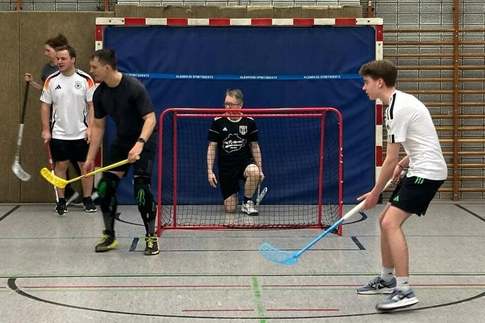 Fünf Personen spielen Floorball in einer Sporthalle, ein Torwart sitzt im Tor, andere halten Schläger in der Hand.