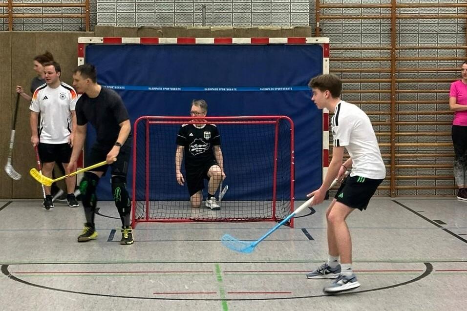 Fünf Personen spielen Floorball in einer Sporthalle, ein Torwart sitzt im Tor, andere halten Schläger in der Hand.