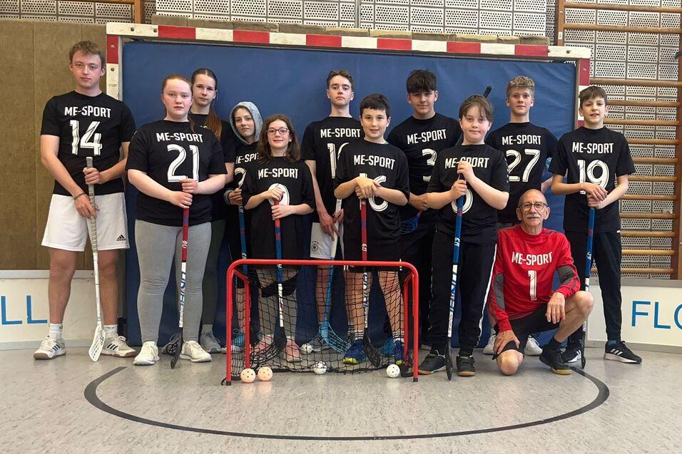 Gruppe von Jugendlichen in Sportshirts mit Bodenballschlägern vor einem roten Tor in einer Sporthalle.