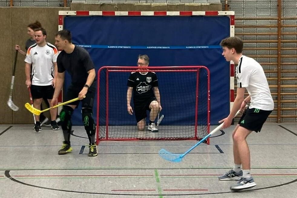 Fünf Personen spielen Floorball in einer Sporthalle, ein Torwart sitzt im Tor, andere halten Schläger in der Hand.