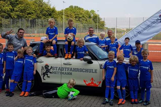Gruppe von Kindern in blauen Fu&szlig;balltrikots posiert fr&ouml;hlich vor einem Auto mit Fahne der Stadtfaahrschule Mettmann.
