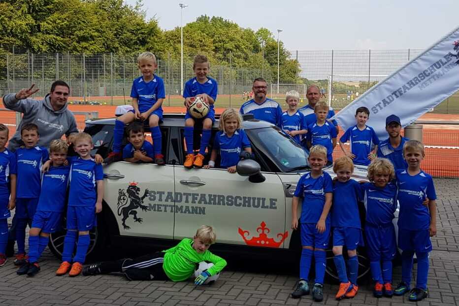 Gruppe von Kindern in blauen Fußballtrikots posiert fröhlich vor einem Auto mit Fahne der Stadtfaahrschule Mettmann.