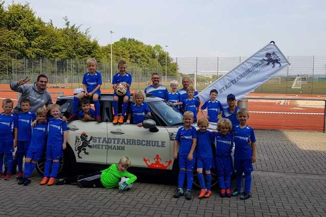 Gruppe von Kindern in blauen Fußballtrikots posiert fröhlich vor einem Auto mit Fahne der Stadtfaahrschule Mettmann.