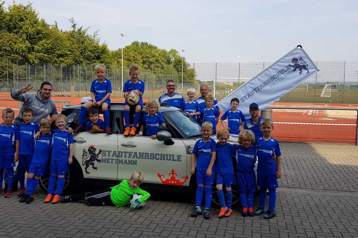 Gruppe von Kindern in blauen Fußballtrikots posiert fröhlich vor einem Auto mit Fahne der Stadtfaahrschule Mettmann.