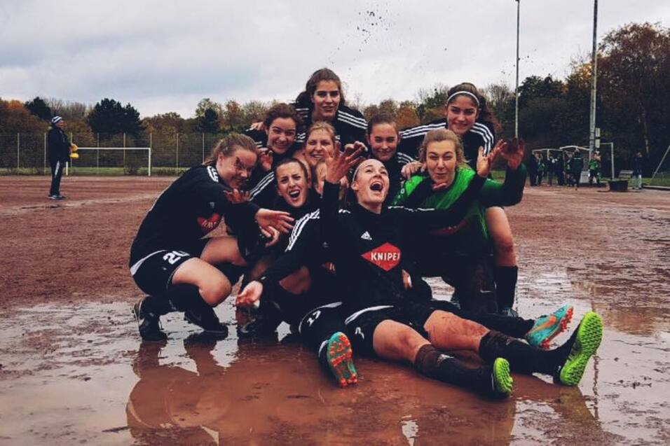Gruppenselfie von neun Frauenfußballspielerinnen, die in einer Pfütze sitzen, lächeln und feiern auf einem Sportplatz.