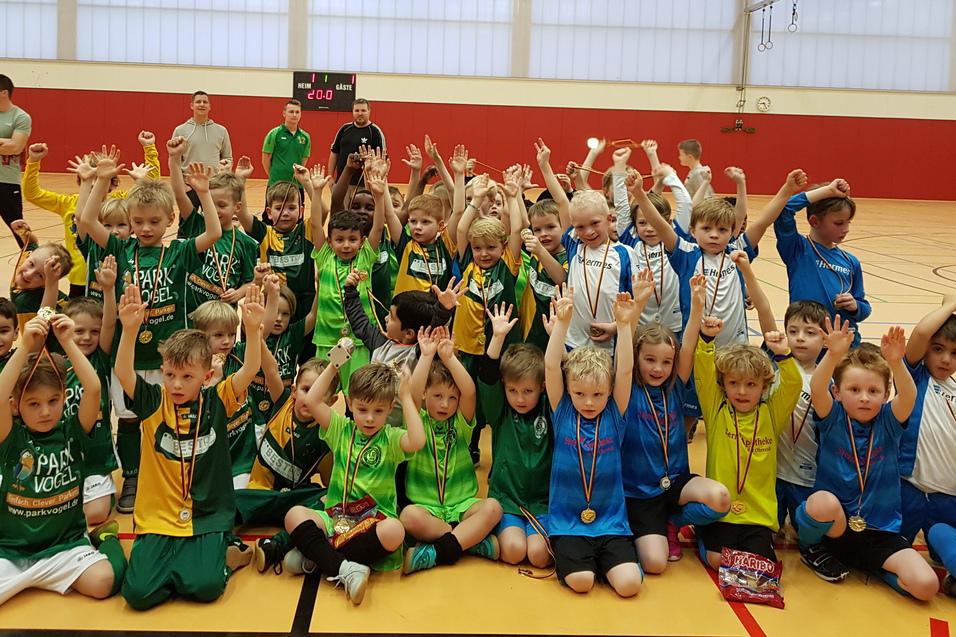 Gruppenshot von zahlreichen Kindern in farbigen Trikots mit Medaillen, die jubelnd in einer Sporthalle stehen.