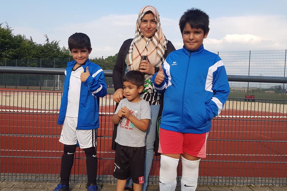 Gruppe von drei Kindern und einer Frau in Sportkleidung, stehend vor einem Fußballfeld mit rotem Kunstrasen.