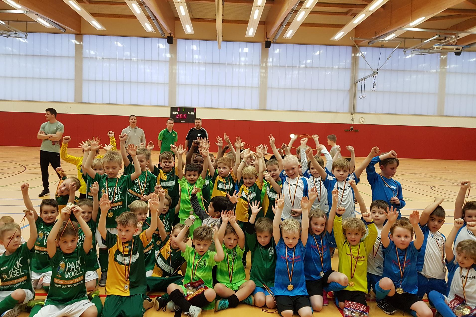 Gruppenshot von zahlreichen Kindern in farbigen Trikots mit Medaillen, die jubelnd in einer Sporthalle stehen.