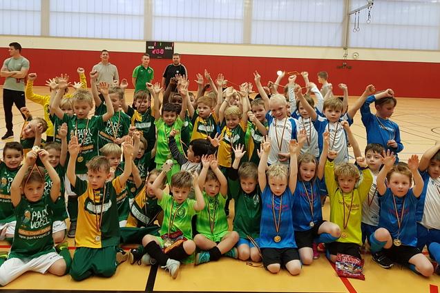 Gruppenshot von zahlreichen Kindern in farbigen Trikots mit Medaillen, die jubelnd in einer Sporthalle stehen.