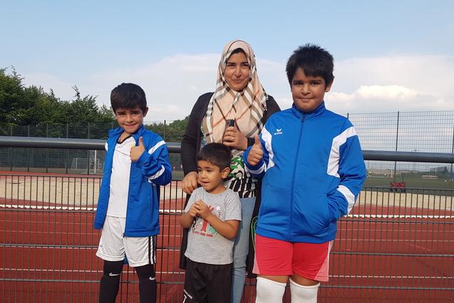 Gruppe von drei Kindern und einer Frau in Sportkleidung, stehend vor einem Fußballfeld mit rotem Kunstrasen.