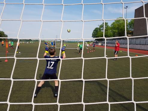 Kind im Tor mit Handschuhen sieht einem Ball entgegen, während Spieler auf dem Fußballfeld trainieren.